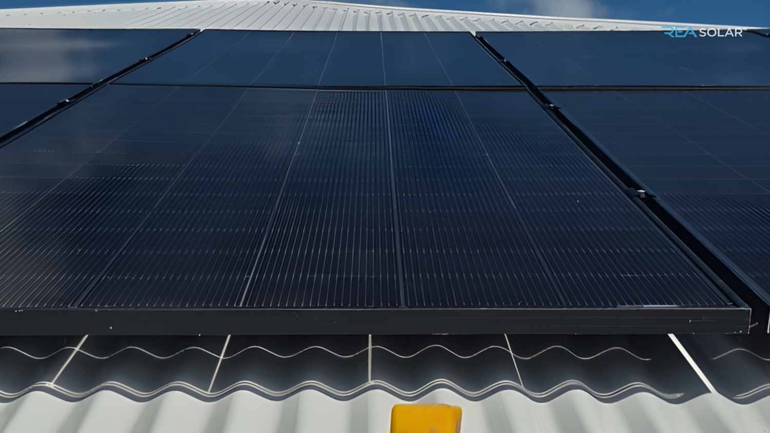 A row of black solar panels is installed on a gray tiled rooftop under a clear blue sky.