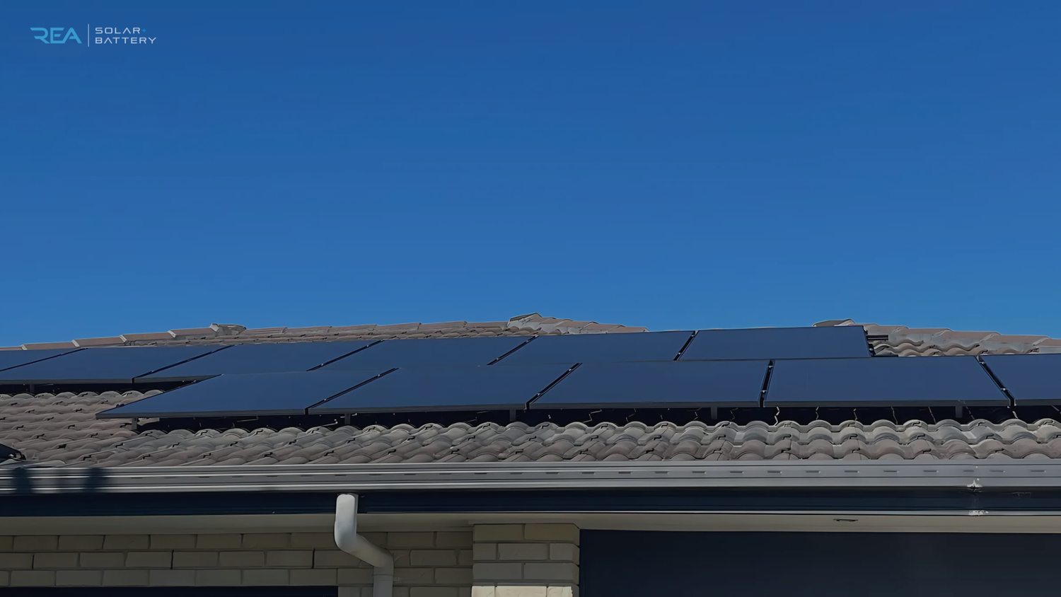 A row of solar panels installed on a sloped, tiled roof under a clear blue sky. The scene conveys a sense of sustainability and clean energy.