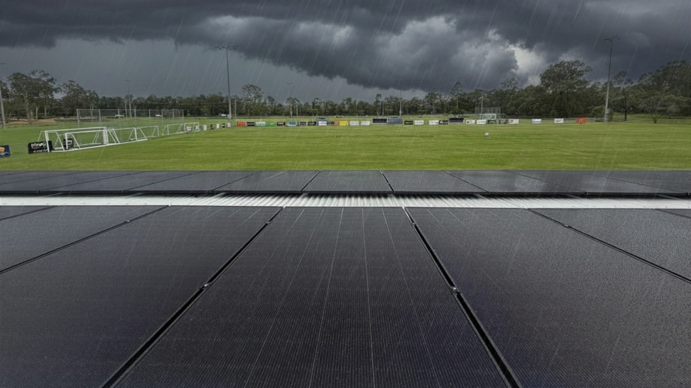 Dark clouds loom over a green sports field, bordered by solar panels in the foreground. The atmosphere is tense and stormy, with impending rain.