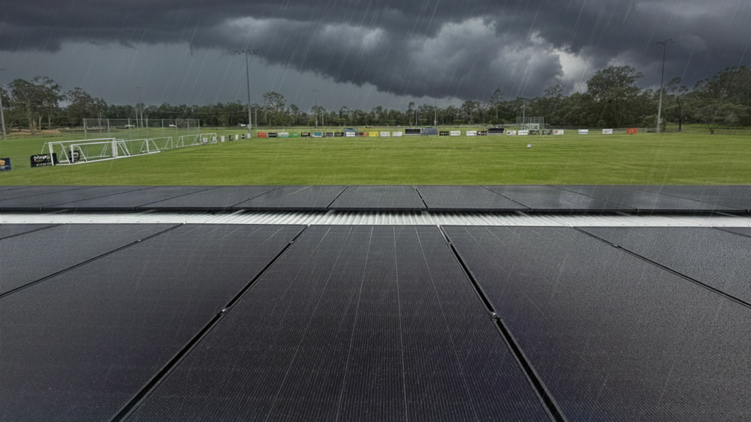 Dark clouds loom over a green sports field, bordered by solar panels in the foreground. The atmosphere is tense and stormy, with impending rain.