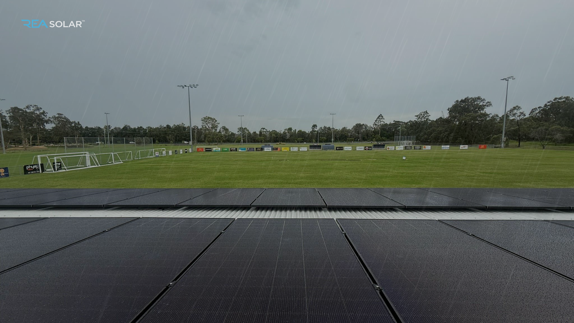 Rain falling on a solar panel installation overlooking a grassy sports field with goalposts. Overcast sky and trees in the background, conveying a calm, rainy atmosphere.